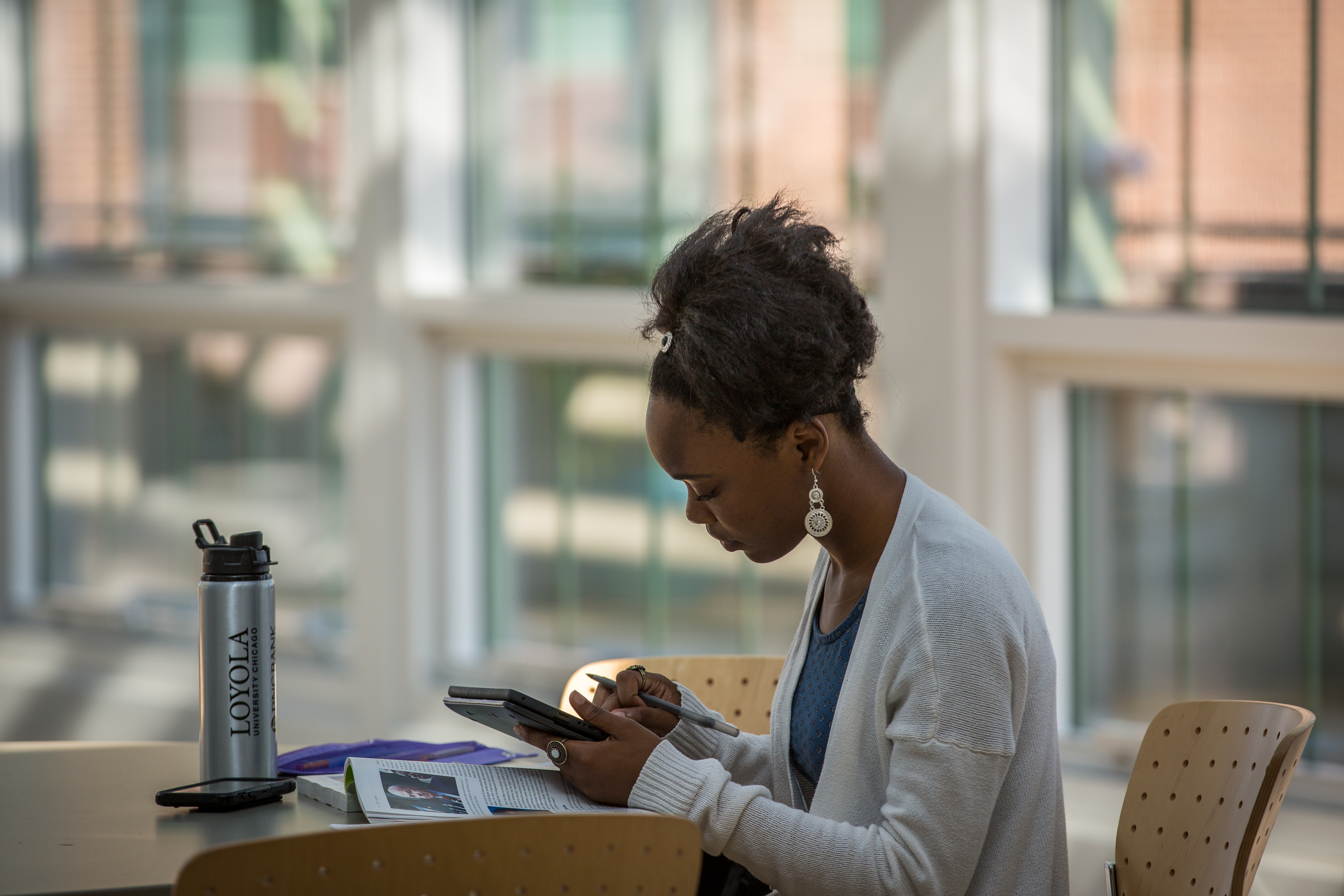 Photo shows student in front of windows looking at tablet with open book on table and Loyola water bottle.